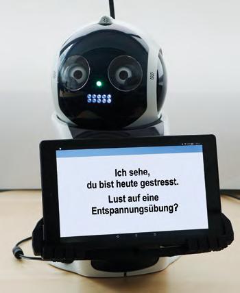 A robot holding a tablet on which is written: Ich sehe, du bist heute gestresst. Lust auf eine Enspannungsübung?